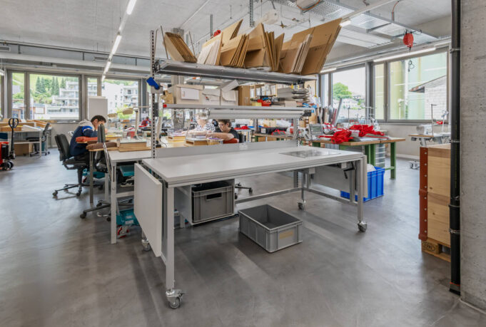 ERGO_line_AG-Packtische-BEWO-Genossenschaft-Oberburg-012 Packtisch ERGO-Basic mit Rollen in einer hellen Werkstatt mit Lagerregalen und Mitarbeitern